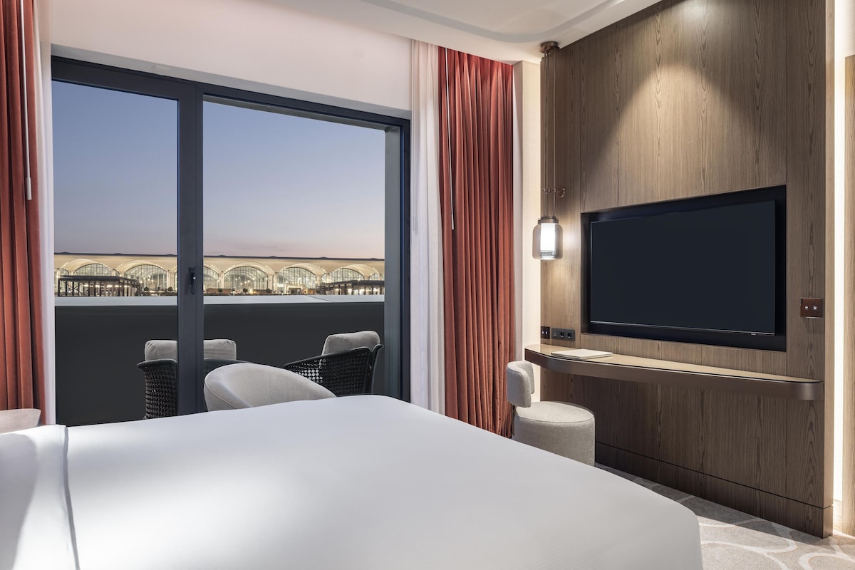 A hotel room with a large bed, a TV mounted on a wooden wall, and a view of a building through the window.