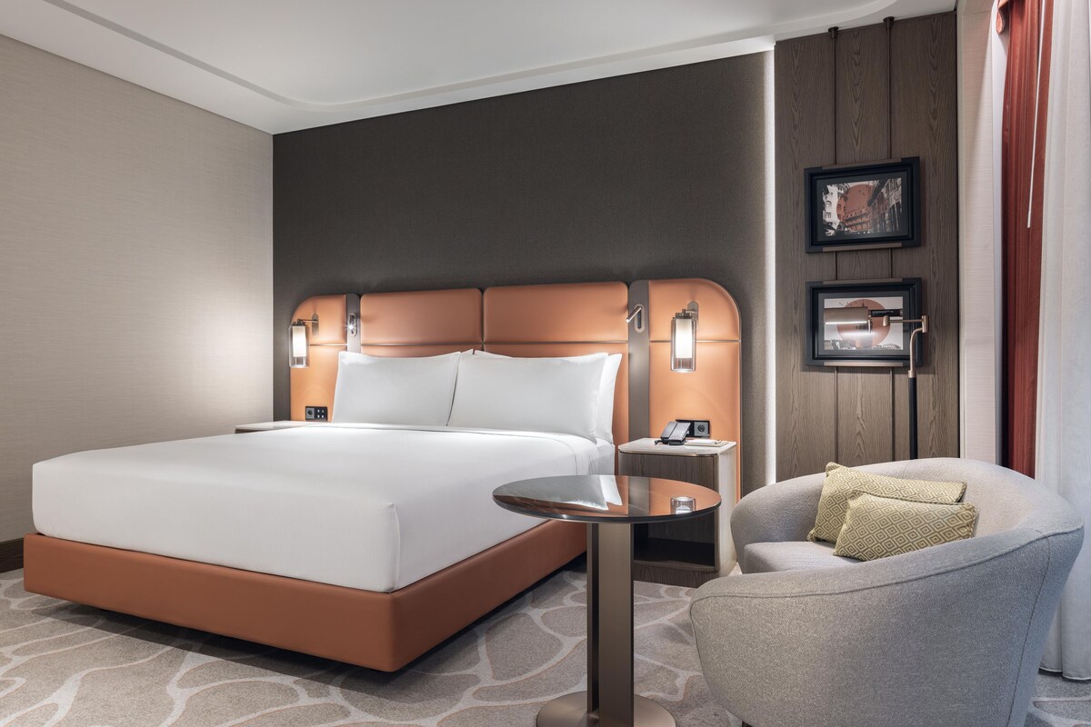 A hotel room with a large bed, a bedside table, a chair, and a small table.