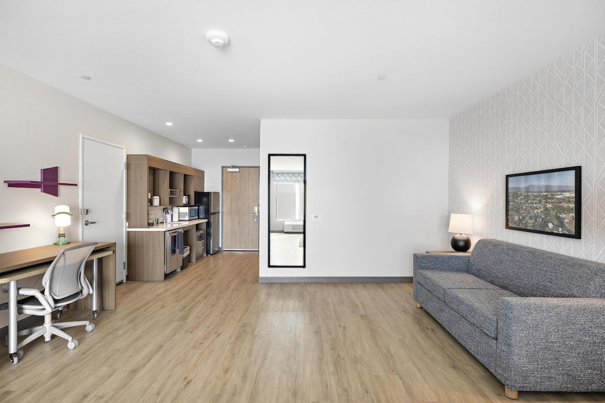 A modern hotel room with a wooden floor, a sofa, a desk with a chair, a kitchenette, and a framed picture on the wall.