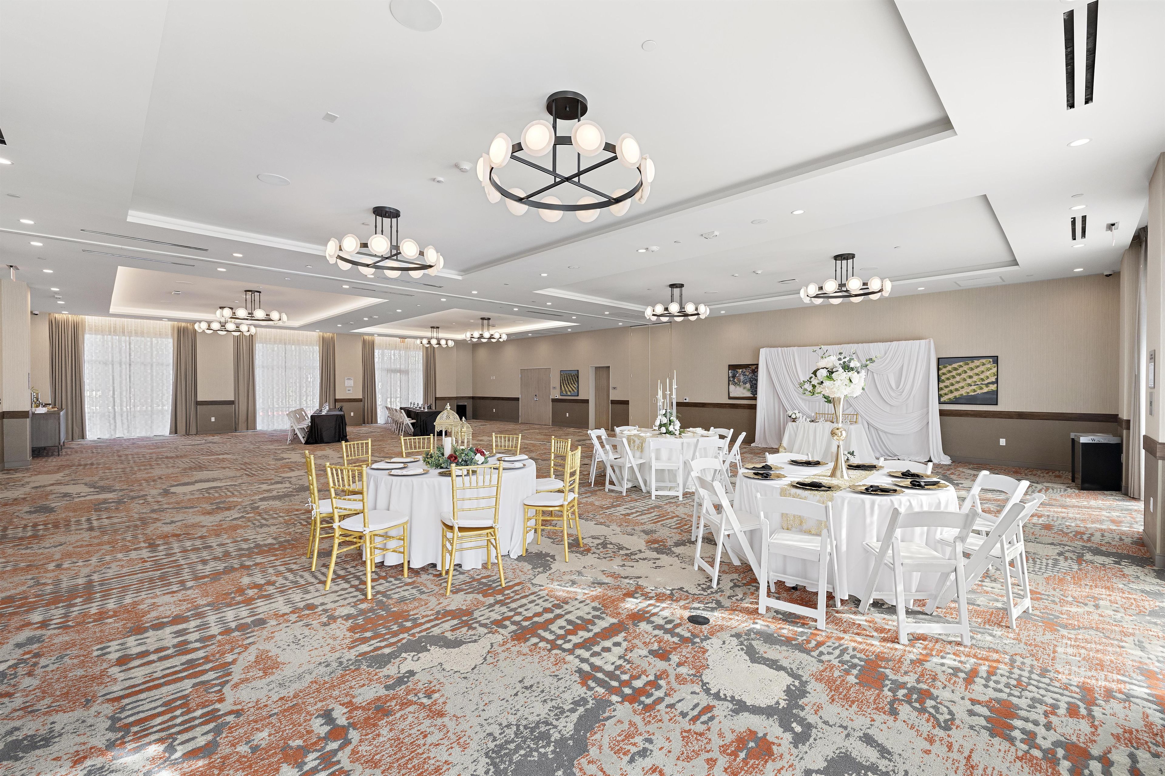 A spacious banquet hall with multiple round tables, white chairs, and elegant chandeliers.