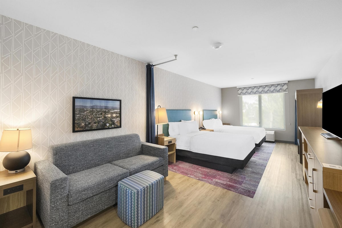A hotel room with a bed, a sofa, a desk, a TV, and a cityscape painting on the wall.