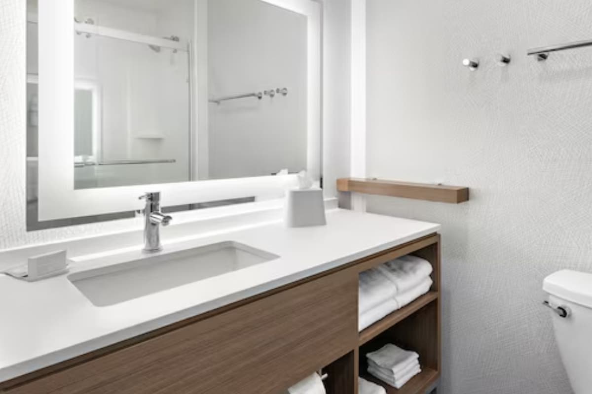 A modern bathroom with a white sink, mirror, and toilet. There are wooden shelves with towels and a towel rack above the toilet.