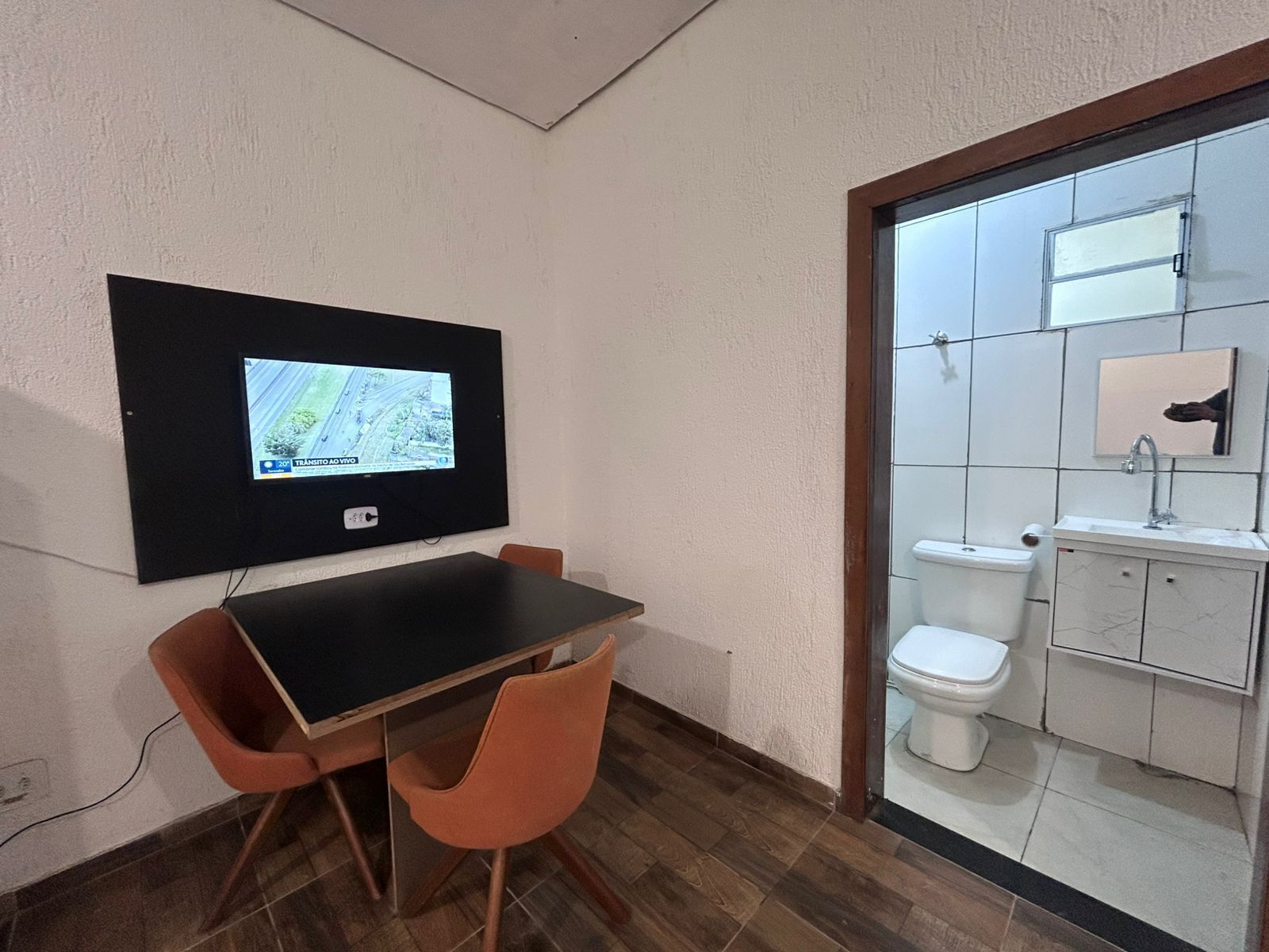 A small room with a wooden table and two chairs, a mounted TV displaying a map, and a bathroom with a toilet and sink.
