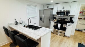 A modern kitchen with white cabinets, a stainless steel refrigerator, a microwave, and a gas stove. There are three black bar stools at the island sink.