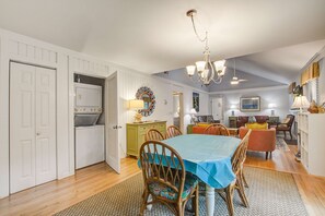 Un comedor con una mesa lista para cuatro personas, rodeada de sillas de madera. Un candelabro cuelga del techo, y se ve una cocina con microondas y estufa al fondo.