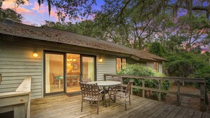 Um deck de madeira com uma mesa, cadeiras, um banco e uma porta de correr de vidro que dá para o interior.