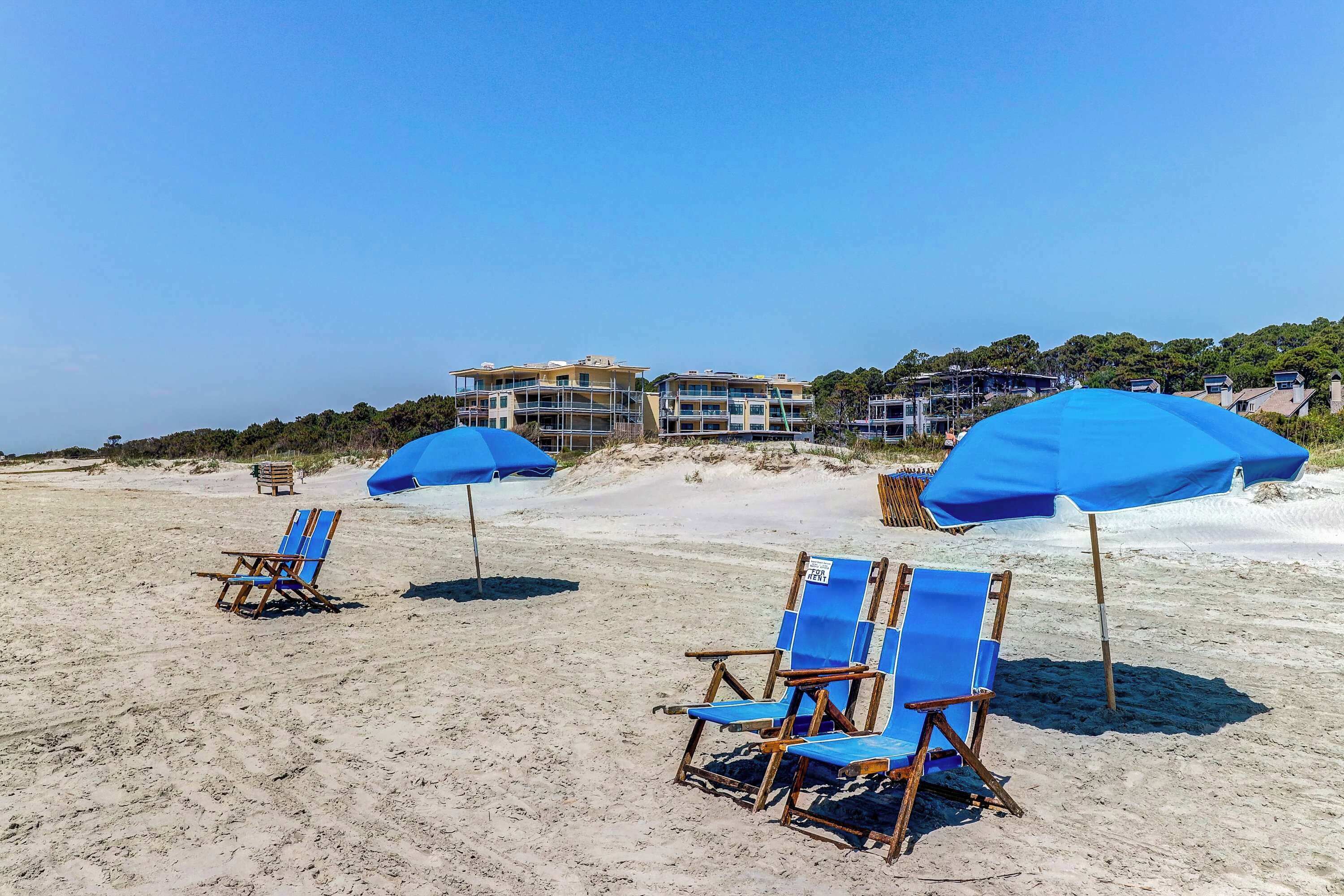 A beach with blue umbrellas and beach chairs, a building in the background, and a clear sky.