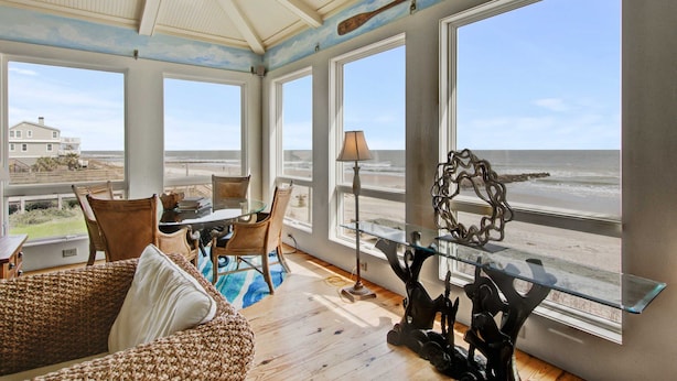 A room with large windows overlooking the beach, a wicker sofa, a glass coffee table, and a dining area with chairs. - Luxury, Dog-Friendly, Oceanfront Home on Folly Beach (Folly Beach)