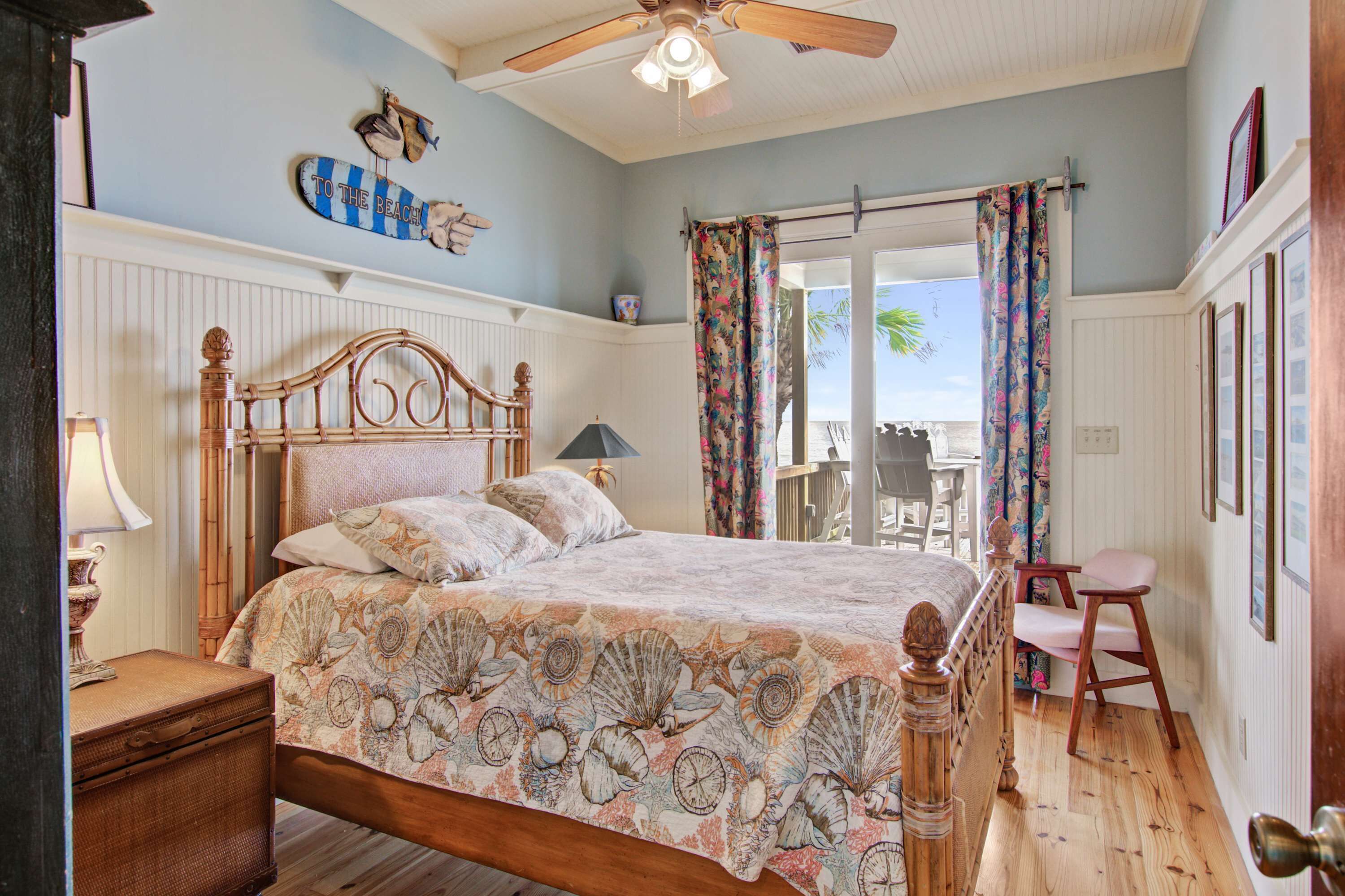 A bedroom with a wooden bed, a ceiling fan, and a view of the beach through a door.