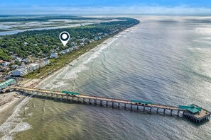 An aerial view of a coastal area with a long pier extending into the water, surrounded by residential buildings and greenery. - Coastal dog-friendly home with pool, ocean views & fenced yard (Folly Beach)