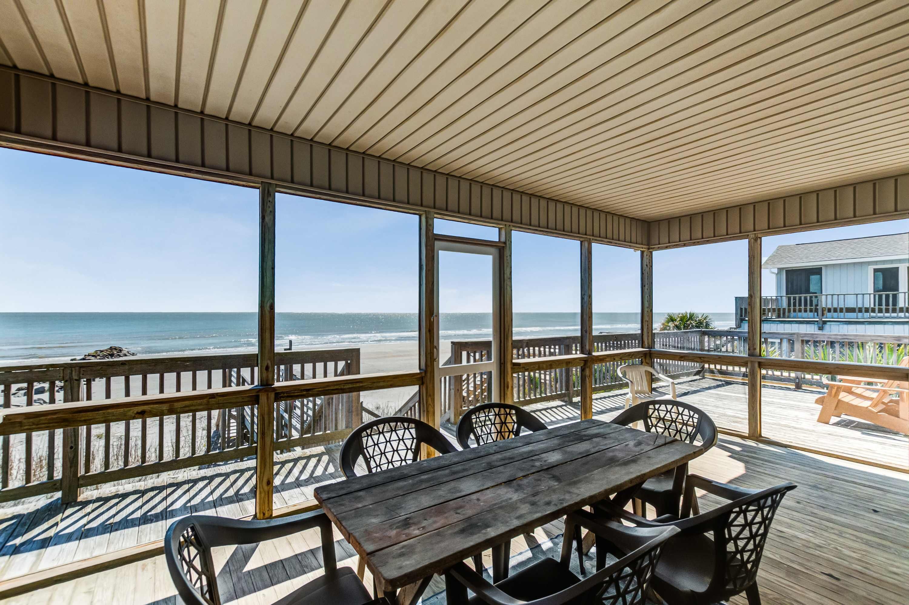 A covered outdoor area with a wooden deck, a table with chairs, and a view of the beach and ocean.