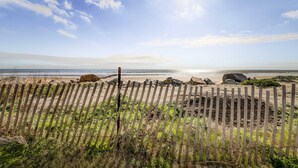 A beach with a wooden fence in the foreground, a rocky shoreline, and a clear sky. - Raised oceanfront home with elevator & outdoor shower just steps from beach (Folly Beach)