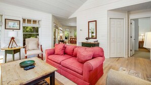 Ein geräumiges Wohnzimmer mit einem roten Sofa, einem Holztisch und einer grünen Schüssel. Dort steht ein Sessel, ein kleiner Tisch mit einer Lampe und eine Tür zu einem weiteren Raum.