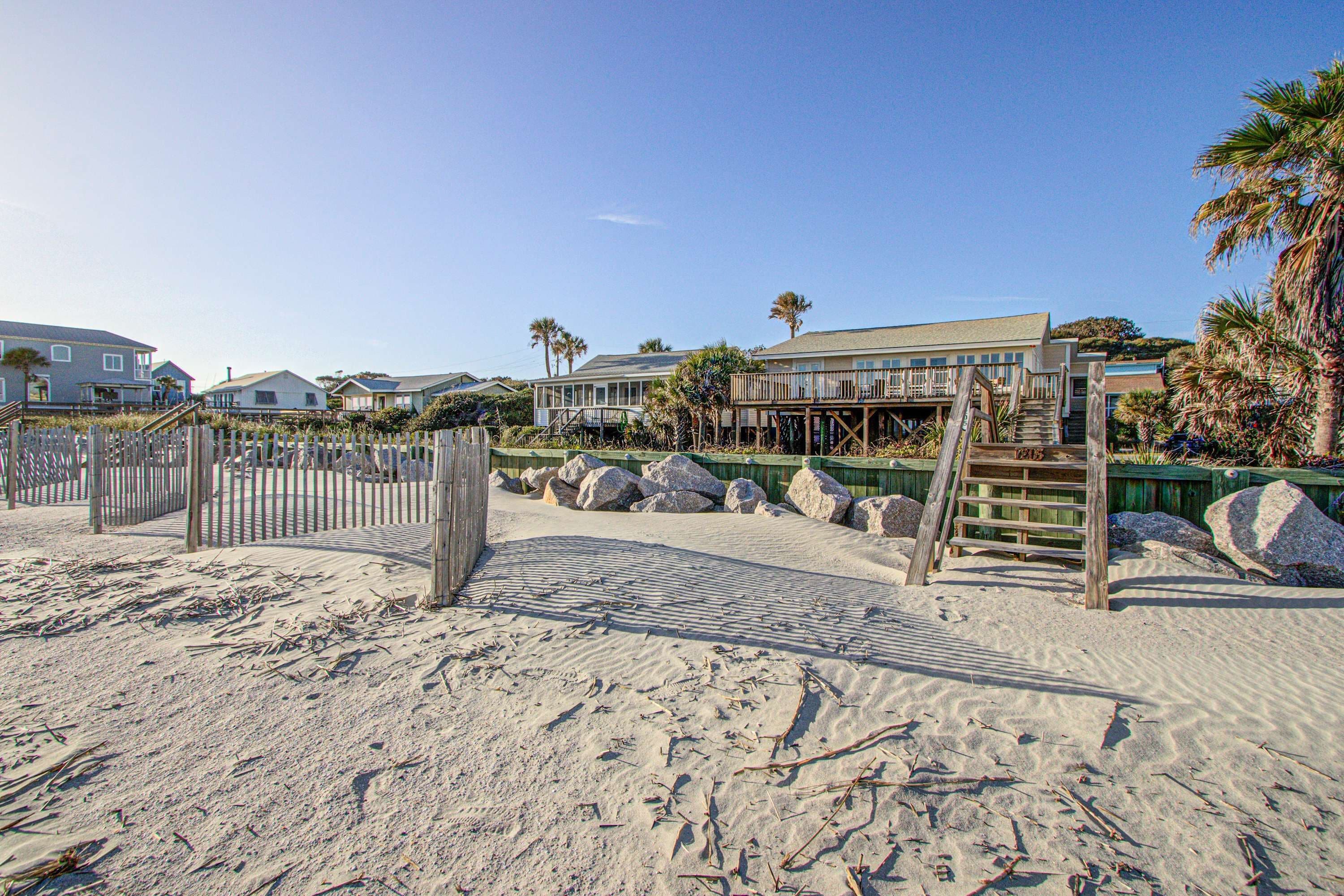 Une propriété située en bord de mer, dotée d’une terrasse en bois, d’un escalier menant à un balcon et d’une plage de sable.