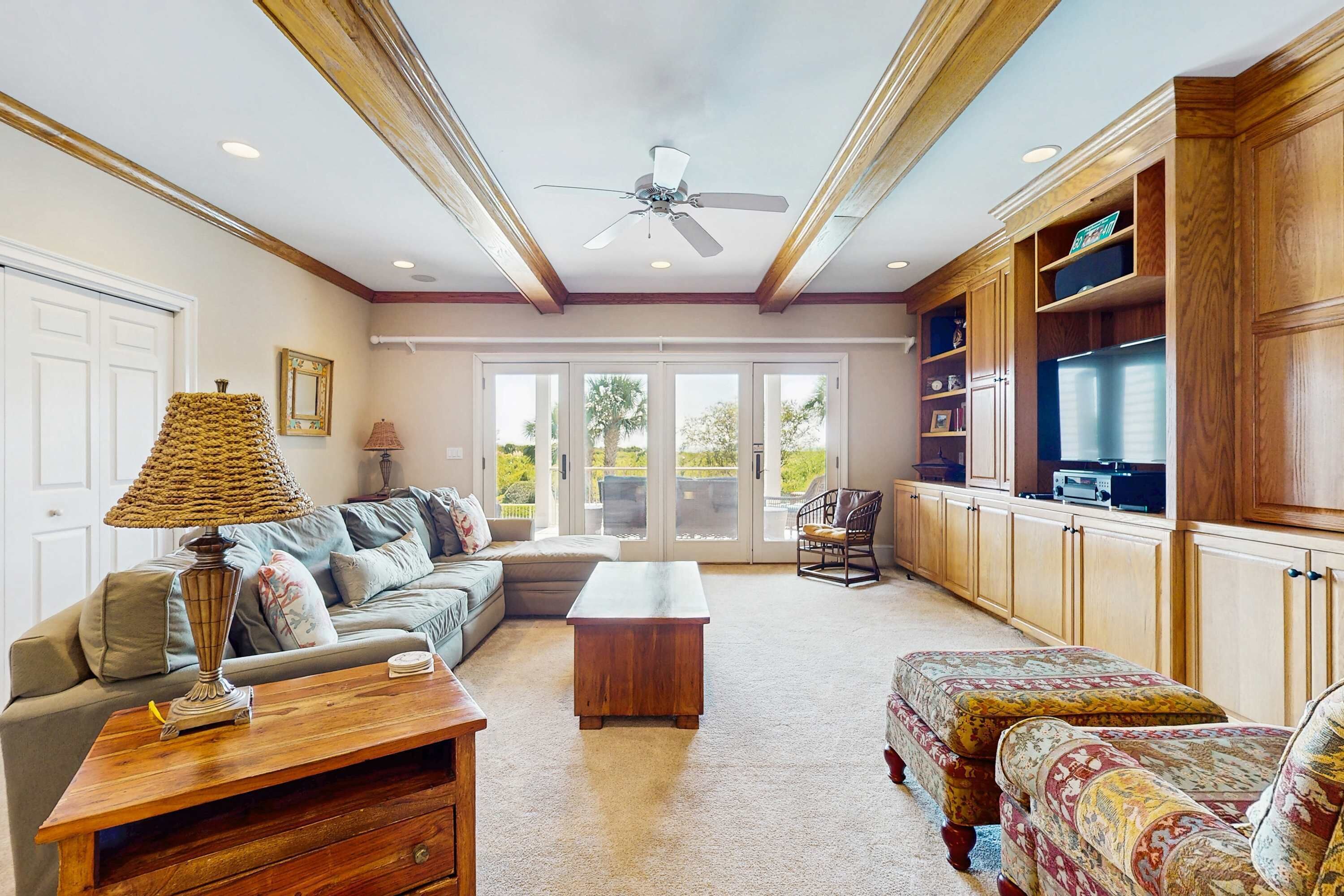A spacious living room with a sofa, a coffee table, and patterned ottomans. There is a television on a wooden cabinet and a ceiling fan.