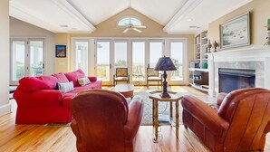A spacious living room with a red sofa, leather armchairs, a fireplace, and large windows. - Gorgeous home with panoramic ocean views, private pool, & hot tub (Seabrook Island)