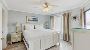 Un dormitorio con cama, mesitas de noche, ventilador de techo, ventana con cortinas y un adorno en la pared.