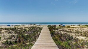Ein hölzerner Steg führt zu einem Strand mit Dünen und spärlicher Vegetation.