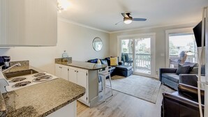 A modern kitchen with granite countertops, white cabinets, and a built-in stove. There is a dining area with a dark-colored sofa and a small table. A large window provides natural light and a view of the outdoors. - Updated Oceanfront Condo w/ Balcony, Pool & Beach Access (Isle of Palms)