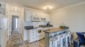 A modern kitchen with white cabinetry, a built-in microwave, a stove, and a bar area with white stools. - Updated Oceanfront Condo w/ Balcony, Pool & Beach Access (Isle of Palms)
