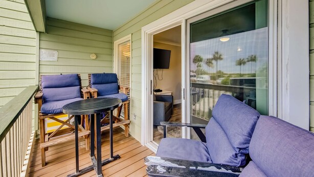 A covered outdoor area with a wooden deck, two blue cushioned chairs, a small round table, and a sliding glass door leading to an interior space. - Updated Oceanfront Condo w/ Balcony, Pool & Beach Access (Isle of Palms)