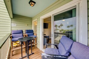 Un espacio exterior techado con terraza de madera, dos sillas azules acolchadas, una mesa redonda pequeña y un ventanal corredero que da a un espacio interior.