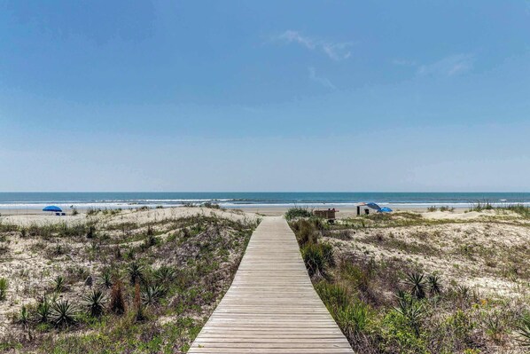 Ein hölzerner Steg führt zu einem Strand mit Dünen und vereinzelter Vegetation.