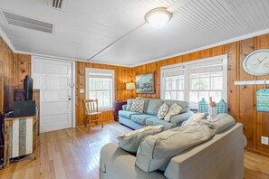 A cozy living room with wooden walls and floors, a blue sofa, a grey sofa, a television on a wooden stand, and a painting on the wall. - Downtown Folly Beach home with free WiFi, washer/dryer, smart TV, & more (Folly Beach)