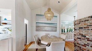 Uma sala de jantar com uma mesa de madeira, duas cadeiras, um lustre e uma pintura grande de uma paisagem marítima.