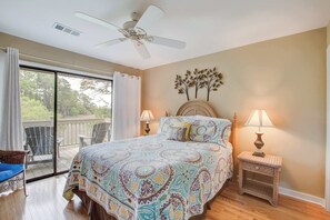 A bedroom with a bed, two bedside lamps, a ceiling fan, a window with curtains, and a view of trees.