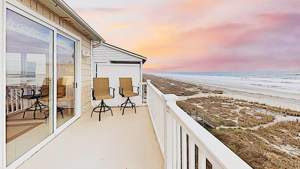 Un balcon avec deux fauteuils en osier et une porte vitrée donnant sur une pièce. Le balcon offre une vue sur une plage avec des dunes et l’océan.