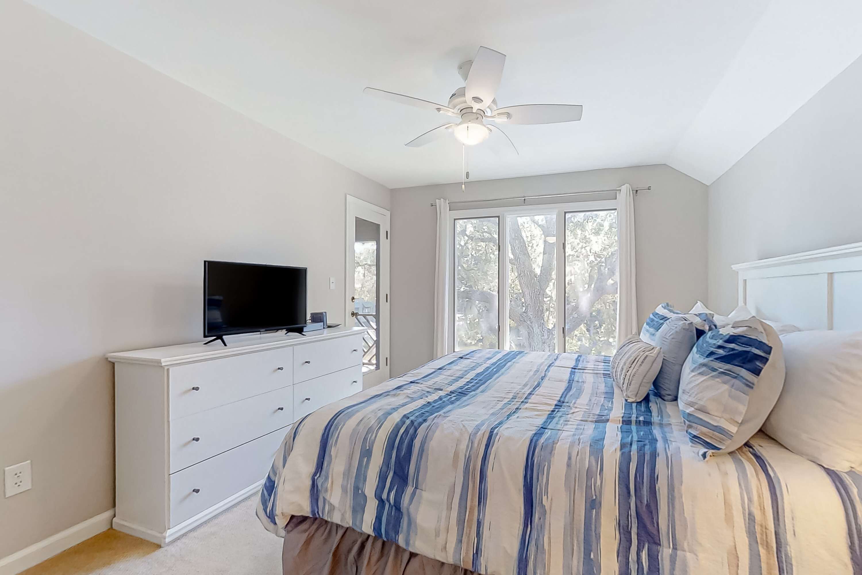 Une chambre à coucher avec un lit, un téléviseur posé sur une commode, un ventilateur de plafond et de grandes fenêtres.