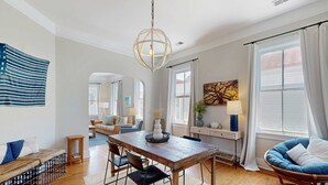 A spacious living room with a wooden dining table, comfortable seating, a large window with curtains, and a hanging light fixture. - Historic upstairs condo in downtown Charleston w/free WiFi - perfect for groups (Charleston)
