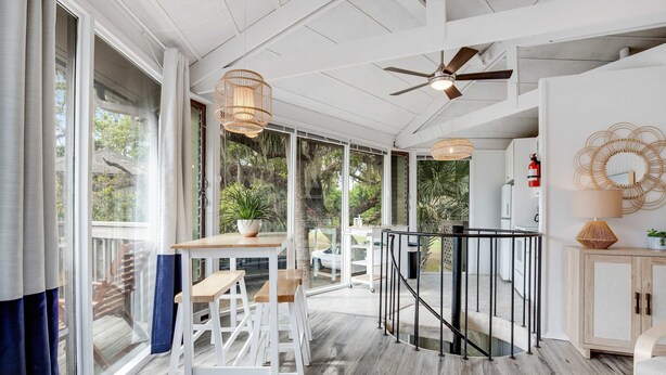 A spacious room with a wooden floor, a dining area with white chairs, a black railing, and large windows offering a view of greenery. - Lakefront home with lagoon view, deck & AC - close to beach, golf, & pools (Seabrook Island)