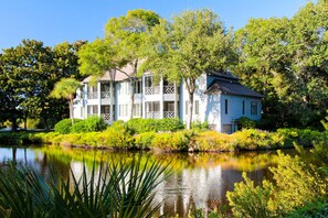A large, two-story house with a balcony, surrounded by lush greenery and a pond.