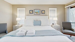 Quarto de hotel com uma cama grande, duas mesas de cabeceira com luminárias, uma cadeira e uma obra de arte na parede.