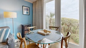 Una stanza con un tavolo in vetro, quattro sedie di legno, un divano con cuscini decorati, una lampada da terra, un quadro incorniciato e vista su alberi attraverso ampie finestre.
