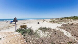 Una spiaggia con una passerella in legno, dune sabbiose e un cielo azzurro limpido.