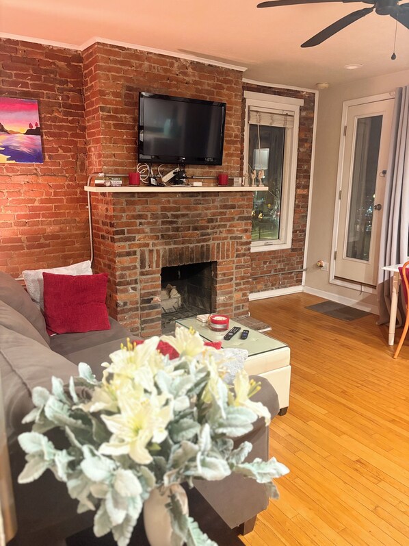 A living room with a brick fireplace, a flat-screen TV mounted above it, a sofa with red cushions, and a glass coffee table with a remote control. - M&T Grandview (Pittsburgh)