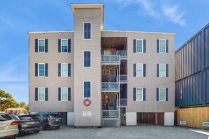 A multi-story building with a central staircase, a sign indicating 'Beachwalk Villas', and a parking area with several cars. - First-floor villa w/partial ocean views, covered porch -one block from the beach (Folly Beach)