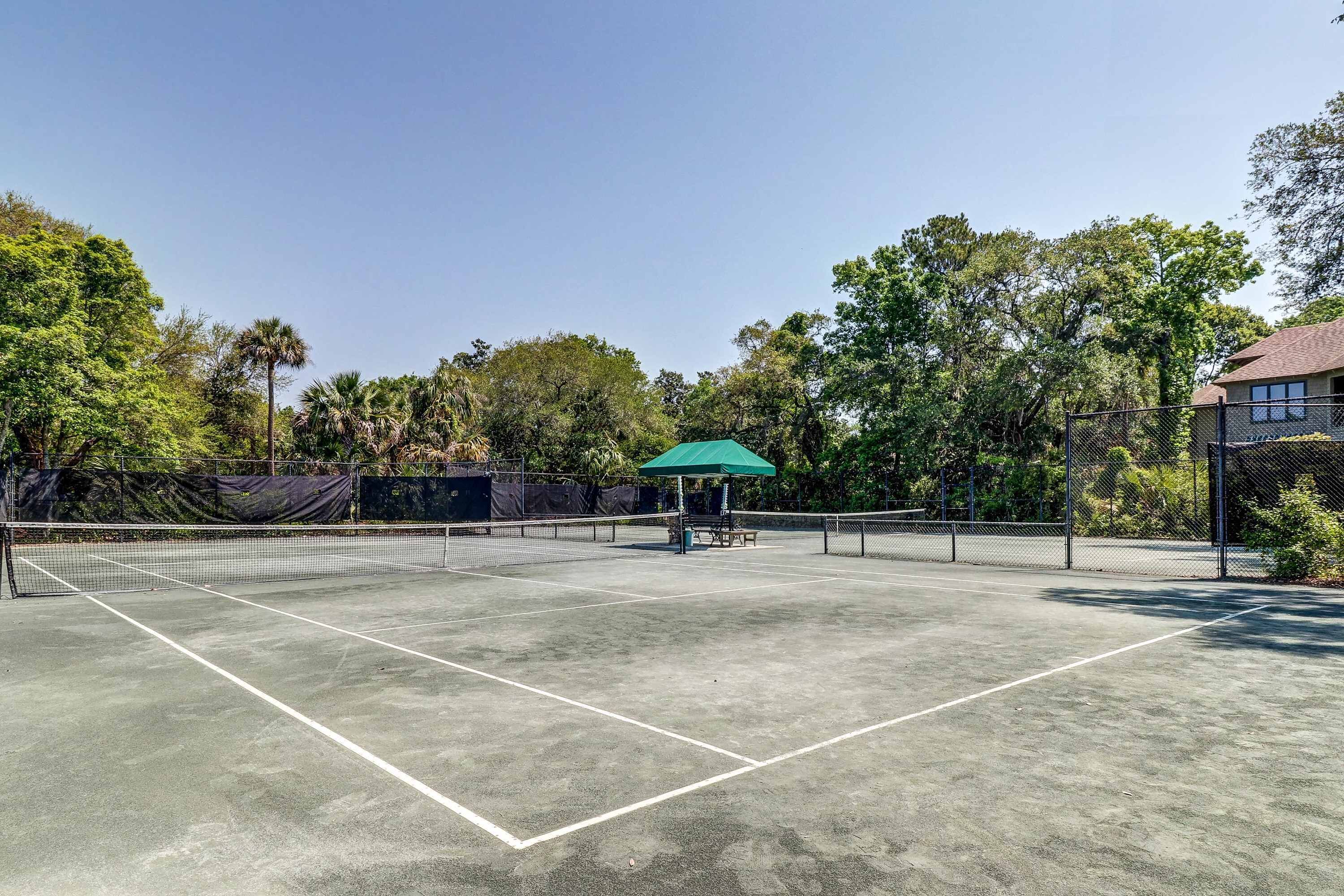 En udendørs tennisbane med net, en grøn parasol og et hegn.