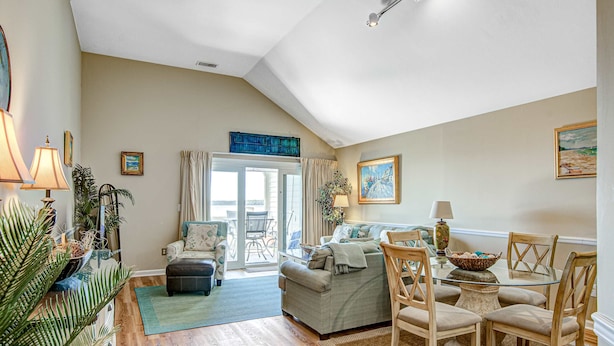 A cozy living room with a sofa, chairs, a glass coffee table, and a dining table. There is a large window with curtains and a view of the outdoors. - Beach villa with full kitchen, ocean/inlet views - short walk to Beach Club (Seabrook Island)