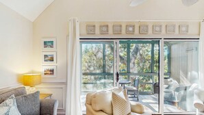 Una sala de estar acogedora con un sofá, una mesa de centro y una lámpara. Hay un ventanal con vistas a árboles y un balcón con mobiliario exterior.