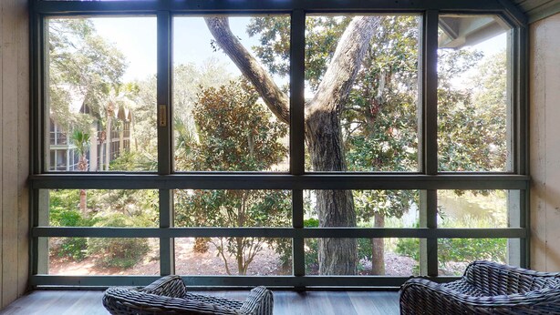 Una habitación con amplios ventanales que ofrecen vistas a árboles y un jardín. Hay dos sillas de mimbre en la habitación.