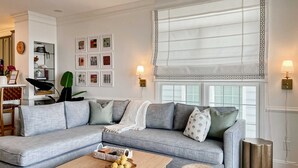 Een woonkamer met een grijze hoekbank, een houten salontafel met fruit en een groot raam met gordijnen met een patroon.