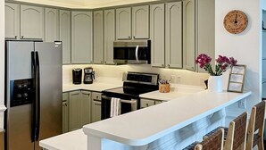 Een moderne keuken met grijze kastjes, roestvrijstalen apparatuur, een wit aanrecht en een decoratieve klok aan de muur.