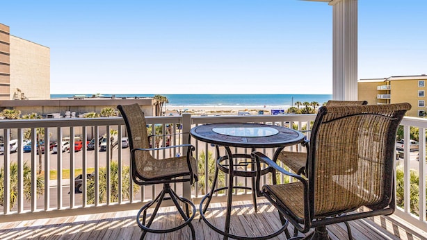A balcony with wicker chairs and a glass table overlooking a beach and ocean.