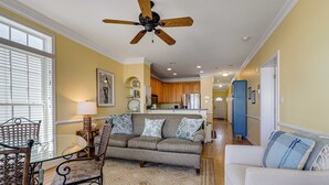 A living room with a sofa, armchair, and glass coffee table. There is a ceiling fan, a lamp, and a window with blinds.
