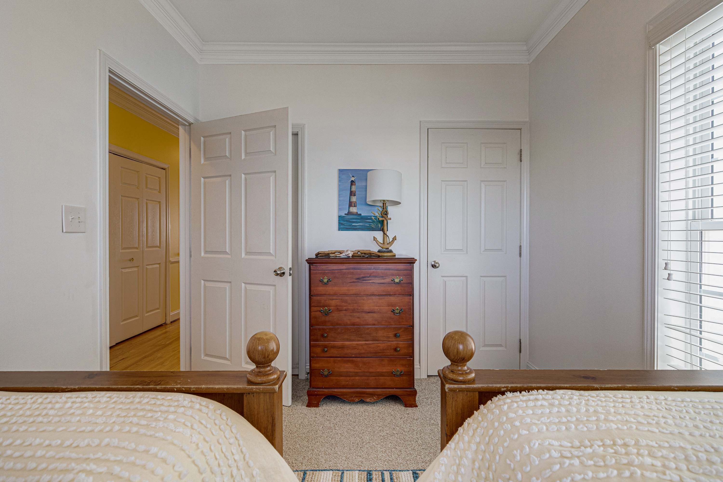 Quarto com cômoda de madeira, duas camas, um quadro na parede e janela com persianas.