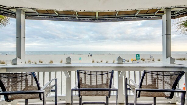 A covered outdoor seating area with wicker chairs, overlooking a beach with people and a lifeguard tower. - Elegant home on the boardwalk with 2 balconies & shared pools - walk to beach (Isle of Palms)